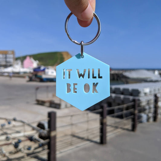 Hexagonal blue keyring with 'IT WILL BE OK' text held by a hand against a blurred coastal background.