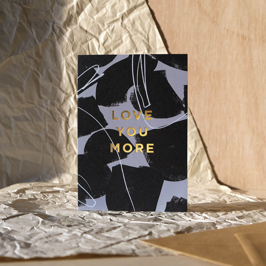 Modern Love Card reads ' Love You More' printed in gold foil on an abstract grey and black background with kraft brown envelope