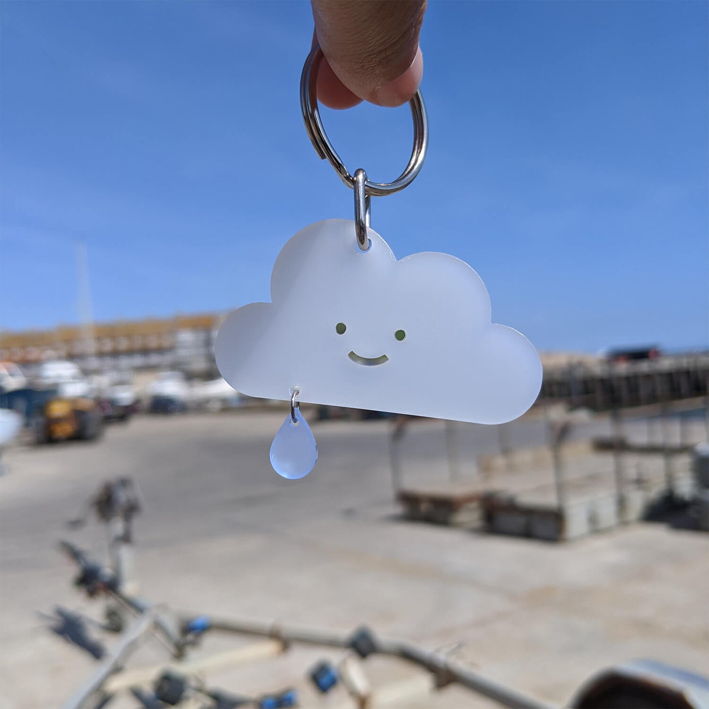 Cloud-shaped keyring with a smiley face and blue raindrop held against a blurred coastal background.