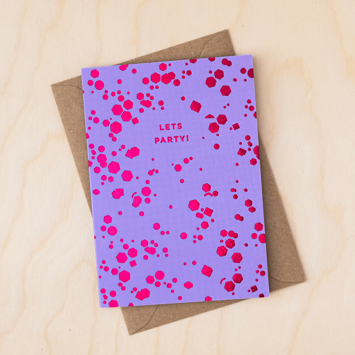 Purple card covered in pink foil confetti shapes and a message that reads Lets Party with a brown kraft envelope