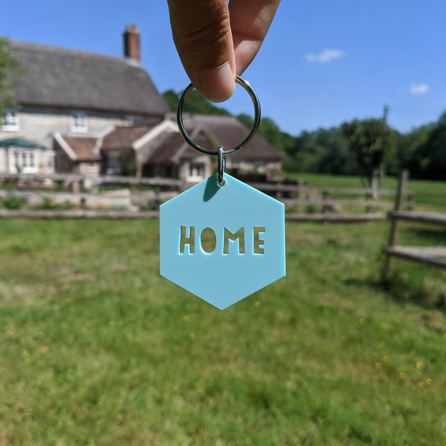 Mint 'HOME' keyring held in front of a countryside scene with a house and fields.