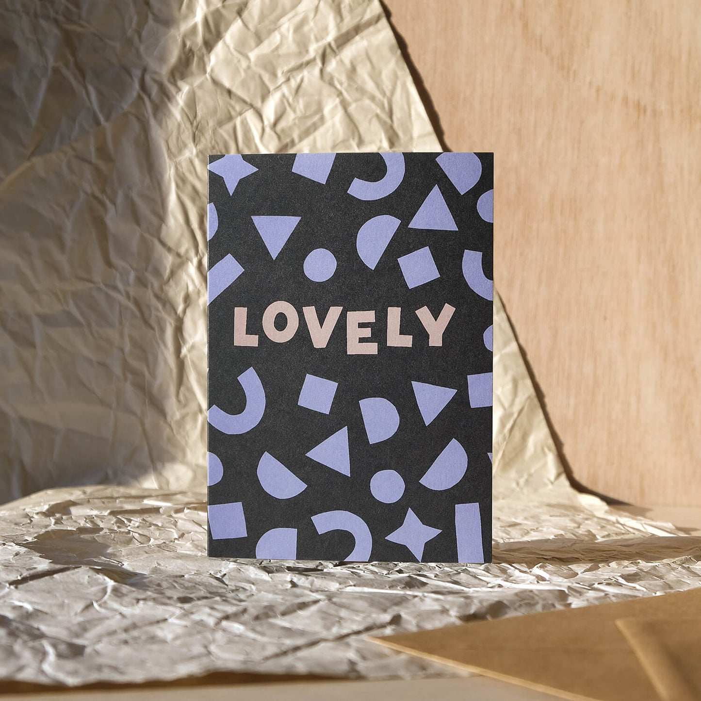 Modern celebrations greetings card featuring bold lilac shapes framing the hand lettered word 'Lovely' on a dark grey background with kraft brown envelope
