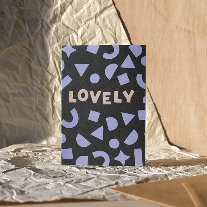 Modern celebrations greetings card featuring bold lilac shapes framing the hand lettered word 'Lovely' on a dark grey background with kraft brown envelope